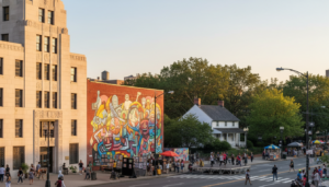 A city street scene featuring a large, colorful abstract mural on a brick wall, situated between a tall Art Deco building and a white colonial-style house. People are active on the street with vendor stalls and an outdoor stage.